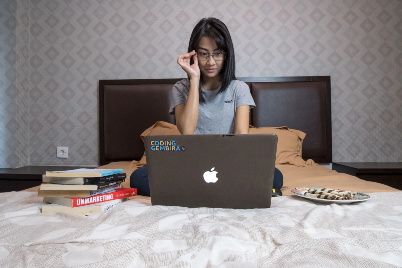 A young woman in glasses sitting on her bed, reading carefully on her laptop